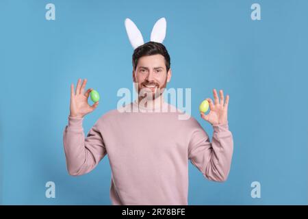 Glücklicher Mann mit niedlichem Hasenohren-Stirnband, der Ostereier auf hellblauem Hintergrund hält Stockfoto