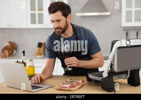 Mann mit einem Glas Wein beim Abendessen, während er sich den Online-Kochkurs mit einem Laptop in der Küche anschaute Stockfoto