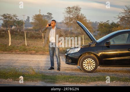 Ein junger Latino in einem Anzug, der vor seinem Auto telefoniert und am Straßenrand eine Panne hat. Stockfoto