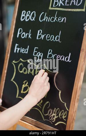 Hand einer jungen Frau, die Street Fast Food verkauft und das Menü mit weißer Kreide zu Beginn des Arbeitstages auf die Tafel schreibt Stockfoto