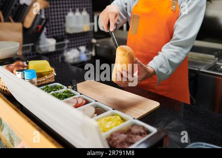 Nahaufnahme eines jungen Lebensmittelverkäufers in einer Orangenschürze, der frische Brötchen über Holzbretter schneidet und Hotdogs für Kunden zubereitet Stockfoto