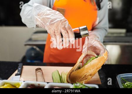Handschuhe von einer jungen Arbeiterin des Street Fast Food Truck, die Soße in einen appetitlichen Hotdog mit einer Vielzahl von Zutaten drinnen gießt Stockfoto