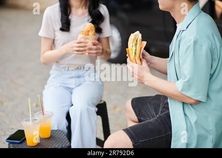 Gekürzte Aufnahme eines jungen Mannes in Freizeitkleidung, der einen appetitlichen Hotdog hat, während er vor seiner Freundin sitzt und einen kleinen Tisch mit zwei Orangensaften hat Stockfoto
