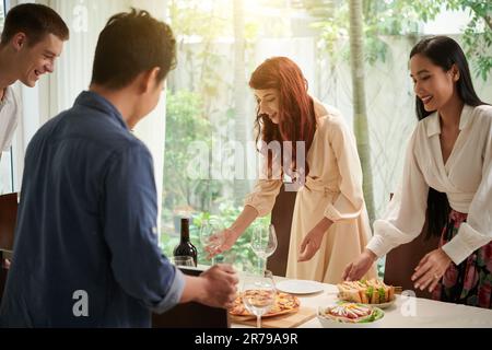Eine Gruppe glücklicher junger interkultureller Freunde serviert am Tisch hausgemachte Pizza, Sandwiches, Flaschen Wein und Weingläser zum Abendessen Stockfoto