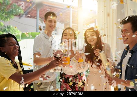 Gruppe junger interkultureller Freunde, die mit Rotwein in Weingläsern und Orangensaft klappern, während sie eine Heimparty mit Konfetti genießen Stockfoto