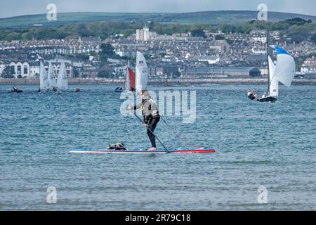 Man paddelt auf dem Paddleboard in Mount's Bay. Kleine Segelboote und die Stadt Marazion im Hintergrund. West Cornwall, England, Großbritannien. Stockfoto