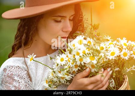 Eine Frau mittleren Alters in einem weißen Kleid und einem braunen Hut hält einen großen Strauß Gänseblümchen in den Händen. Wildflowers für Glückwünsche Stockfoto