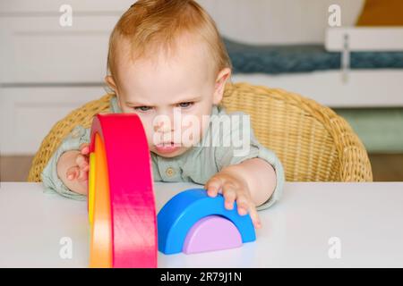 Kleines Baby in Kleidung aus natürlichem Stoff spielt mit regenbogenfarbenen Holzspielzeugen am weißen Tisch. Gemütliche Zimmer im skandinavischen Stil. Stockfoto