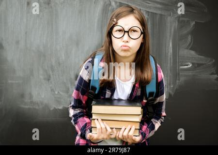 Teenager, kleine Schülerin, die einen Haufen schwerer Bücher auf dem Hintergrund einer Tafel hält. Teenager-Mädchen im Hemd, Brille mit blauem Rucksack und übergewichtigem Stapel Stockfoto