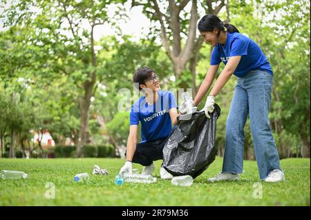 Lächelnde und glückliche junge asiatische Freiwillige mit einem Plastikmülleimer, die gemeinsam den öffentlichen Park aufräumen. Konzept der Freiwilligenarbeit Stockfoto