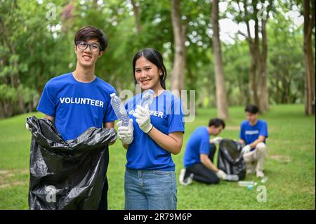 Ein Porträt glücklicher und attraktiver junger asiatischer Freiwilliger in Uniform, der im öffentlichen Park mit einem Müllsack und einer Plastikflasche stand Stockfoto