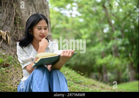 Eine schöne junge Asiatin konzentriert sich darauf, ein Buch zu lesen, während sie am Wochenende auf dem Gras unter einem Baum in einem öffentlichen Park sitzt. Stockfoto