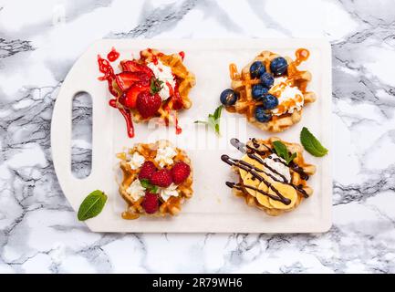 Köstliche belgische Waffeln mit Beere, Obst, Schlagsahne und verschiedenen süßen Belag auf weißem Marmor. Draufsicht. Stockfoto