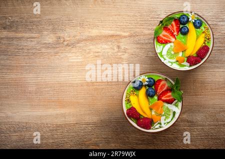 Grüner Smoothie in Schüsseln für gesundes Frühstück. Frischer Bio-Smoothie aus Spinat, Banane, Spirulina, Weizengras und Limette mit Beeren und Obst Stockfoto
