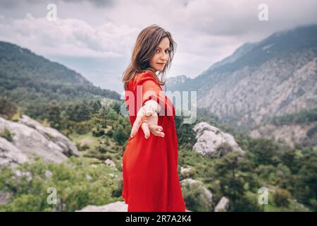 Folgen Sie mir - ein Mädchen in einem roten Kleid auf einer Klippe, das den Blick auf eine Bergschlucht bewundert Stockfoto