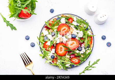 Erdbeersalat mit Rucola, Kopfsalat, Blaubeeren, Fetakäse und Walnüssen, weißer Tisch. Frisches, nützliches Gericht für gesunde Ernährung Stockfoto