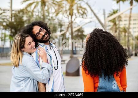 Ein hübsches junges Paar, das mit einem Freund auf der Straße lacht. Drei verschiedene Menschen lächeln, haben Spaß und schauen sich draußen an. Stockfoto