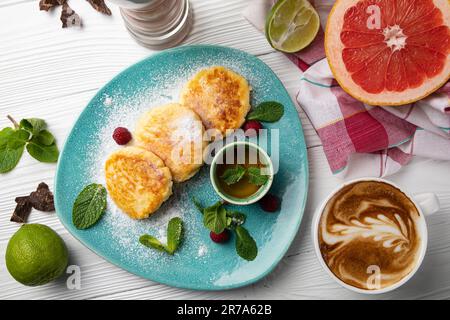 Ein weißer Teller mit einer Vielzahl frischer, reifer Früchte wie Orangen, Bananen und Trauben neben zwei dampfenden Tassen Kaffee auf einem Holztisch Stockfoto