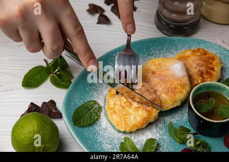 Ein Bild eines Frühstücks bestehend aus einem blauen Teller mit frisch zubereiteten Pfannkuchen und einer Tasse dampfendem Kaffee, wobei eine Person ein Gebäck zum Genießen verwendet Stockfoto