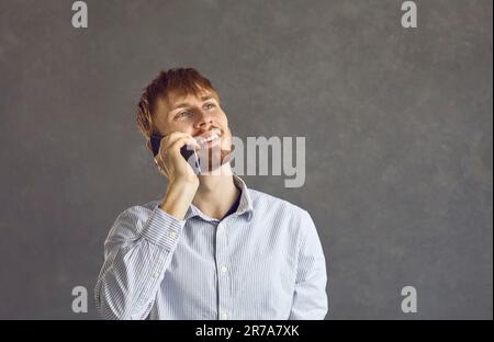 Positiv lächelnder Mann, der mit dem Handy spricht und über grauem Hintergrund steht Stockfoto