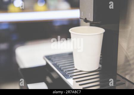 Ein weißer Pappbecher steht auf dem Stand der Kaffeemaschine, gemütliche Atmosphäre eines Cafés, selektiver Soft Focus Stockfoto