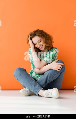 Eine lange, stylische junge Frau mit rotem Kopf in Bluse mit Blumenmuster und Jeans, die auf orangefarbenem Hintergrund lächeln, trendig-lässiger Sommer Stockfoto