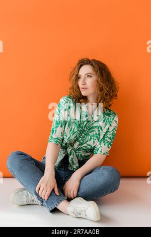 Selbstbewusste junge rothaarige Frau in moderner Bluse mit Blumenmuster und Jeans, die auf orangefarbenem Hintergrund sitzen und wegschauen. Trendig und lässig im Sommer Stockfoto