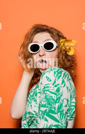 Schockierte junge rothaarige Frau mit Orchideenblume und Sonnenbrille posieren in Bluse mit Blumenmuster und stehen auf orangefarbenem Hintergrund, Sommer-Freizeit Stockfoto