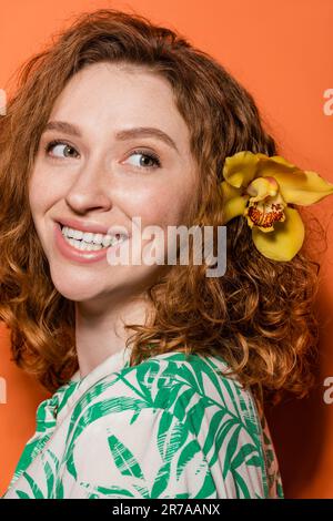 Porträt einer fröhlichen jungen und rothaarigen Frau mit Orchideenblüte im Haar, die wegsieht und in Bluse posiert, während sie im Sommer auf orangefarbenem Hintergrund steht Stockfoto