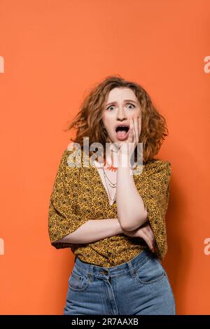 Schockierte junge rothaarige Frau in Bluse mit abstraktem Muster und Jeans, die auf orangefarbenem Hintergrund in die Kamera schauen, stilvolles, legeres Outfit und s Stockfoto