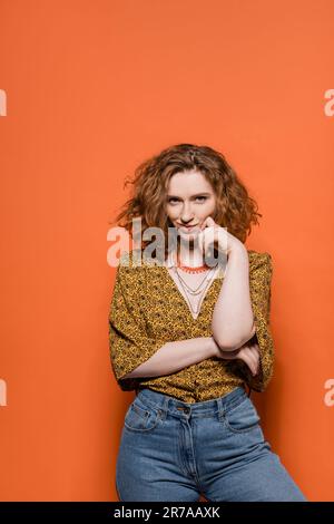 Selbstbewusste und lächelnde rothaarige Frau in Bluse mit abstraktem Muster und Jeans, die auf orangefarbenem Hintergrund auf die Kamera blickt, stilvoll leger Stockfoto