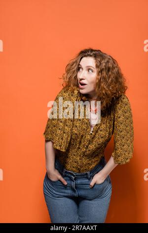 Schockierte junge rothaarige Frau in gelber Bluse und trendige Jeans, die wegblickt, während sie posiert und auf orangefarbenem Hintergrund steht, stilvolles, legeres Outfit Stockfoto