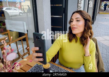 Vorderansicht einer jungen, wunderschönen Latina-Frau argentinischer Herkunft, die ein Foto mit ihrem Handy macht, während sie vor dem Restaurant sitzt, Kopierraum. Stockfoto