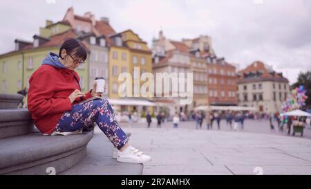 Eine ältere Dame sitzt auf den Stufen und trinkt Kaffee und benutzt ein Smartphone im historischen Zentrum einer alten europäischen Stadt. Palastplatz, Warschauer Altstadt Stockfoto