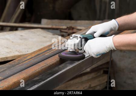 Ein Mann, der mit einem elektrischen Winkelschleifer arbeitet. Entfernen von Rost ein quadratisches Metallrohr. Die Hände in weißen Handschuhen halten ein Elektrowerkzeug, eine Drahtbürste. Stockfoto