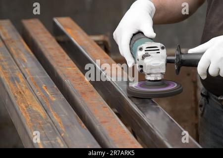 Ein Mann, der mit einem elektrischen Winkelschleifer arbeitet. Entfernen von Rost ein quadratisches Metallrohr. Die Hände in weißen Handschuhen halten ein Elektrowerkzeug, eine Drahtbürste. Stockfoto