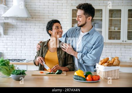 Multirassisches glückliches Paar in lässiger Kleidung, junge Ehepartner, stehen zu Hause in der Küche, bereiten Salat zusammen zu, Frau schneidet Gemüse, Mann umarmt sie, sie sehen sich liebevoll an, lächeln Stockfoto