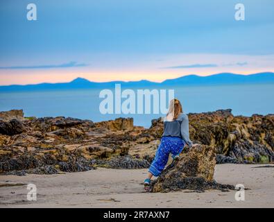 Eine junge Frau sitzt auf einem großen Felsbrocken und blickt an einem sonnigen Tag über ein riesiges Gewässer Stockfoto