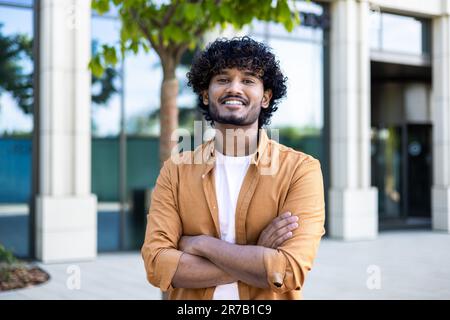 Porträt eines jungen hispanischen Freiberufler-Geschäftsmanns außerhalb des Bürogebäudes, erfolgreicher Mann, der in die Kamera schaut und lächelt und zerknitterte Hände hat, Arbeiter mit lockigem Haar und Hemd. Stockfoto