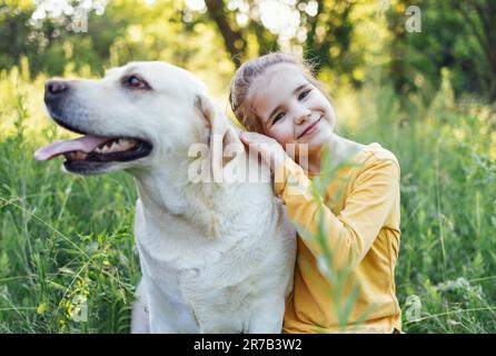 Süßes kleines Mädchen, das ihren Hund umarmt - Golden labrador Retriever. Das süße Baby lächelt und schaut in die Kamera. Hübsches Kind und ihr Haustier draußen in einem Park Stockfoto