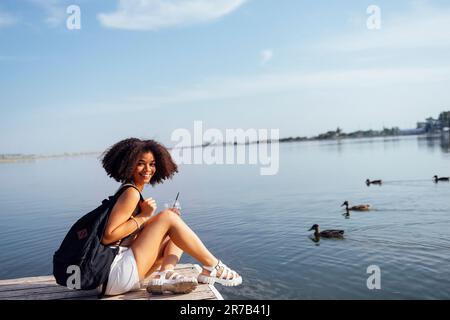 Eine wunderschöne Teenagerin mit gemischter Rasse sitzt auf einem Holzpier und trinkt einen Cocktail in Glas. Charmantes afrikanisches Mädchen in lässiger Kleidung und schwarz Stockfoto