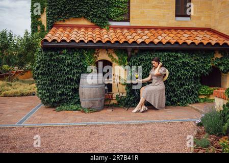 Eine junge Frau in elegantem Kleid und Strohhut sitzt am Tisch auf der Terrasse des Hauses. Hübsches Mädchen mit einem Glas Eisorangen-Cocktail im Garten. Backsteinmauern Stockfoto