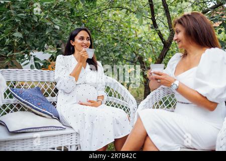 Zwei Frauen halten Tassen in den Händen und trinken köstlichen Tee im Garten. Schwangere junge Frau und ihre Mutter mittleren Alters sitzen auf weißen Korbstühlen und Stockfoto