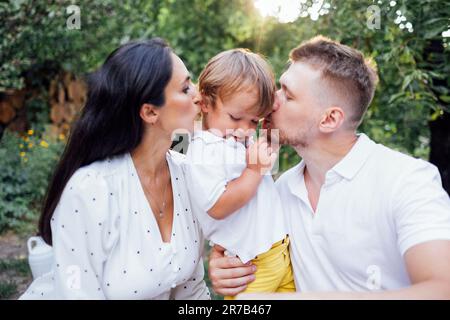 Süße Familie in leichter, lässiger Kleidung im Garten auf der Terrasse des Hauses. Ein junges Paar und ihr Sohn draußen an einem sonnigen Sommertag. Dad hält sich zurück und wartet Stockfoto