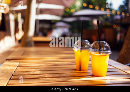 Zwei orangefarbene Cocktails in Plastikgläsern mit Strohhalmen vor der Kulisse eines Open-Air-Cafés. Kalte Limonaden mit Zitronenscheiben auf einem Holztisch Stockfoto