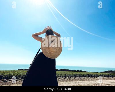 Eine junge Frau in schwarzem, langem Kleid und einem Strohhut mit breiter Krempe steht in anmutiger Pose und schaut auf den Weinberg. Schlankes Mädchen tritt zurück und die Hände auf ihn Stockfoto