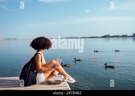 Eine wunderschöne Teenagerin mit gemischter Rasse sitzt auf einem Holzpier und trinkt einen Cocktail in Glas. Charmantes afrikanisches Mädchen in lässiger Kleidung und schwarz Stockfoto