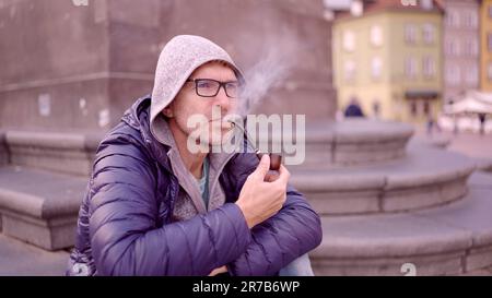 Warschau, Polen. 11. Juni 2023. Erwachsener Mann mit Brille, der auf dem Platz sitzt und eine Tabakpfeife raucht, der Rauch auf dem Palastplatz freisetzt, Warschauer Altstadt (Kreditbild: © Andrey Nekrasov/ZUMA Press Wire), NUR REDAKTIONELLER GEBRAUCH! Nicht für den kommerziellen GEBRAUCH! Stockfoto