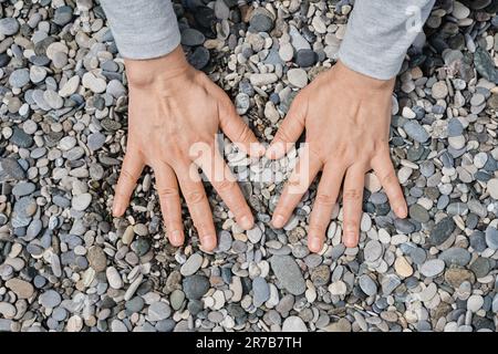 Frauenhände liegen auf den Kieselsteinen. Kieselsteinstrand aus nächster Nähe, dunkle runde Kieselsteine und graue trockene Kieselsteine, hochwertige Fotoidee für die Leinwand Stockfoto