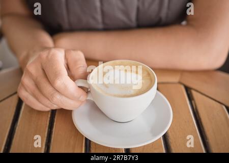 Nahaufnahme einer Tasse duftenden Cappuccino auf dem Tisch in den Händen einer Frau, belebender duftender Kaffee während einer Pause. Selektiver Fokus auf die Finger Stockfoto